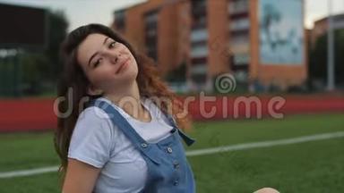 一幅美丽的黑发女人的肖像，穿着蓝色的阳光，坐在体育场的一个足球球门附近的草坪上。 阳光照射
